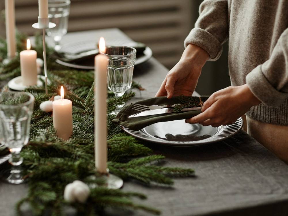 Person som setter et svart tallerken på et dekket bord med lys og grønne dekorasjoner, klar for middagsservering.
