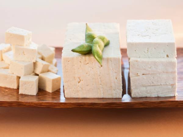 Små terninger av tofu plassert på en treplate med noen grønne bønner på toppen av en blokk tofu, og flere tofu-blokker i bakgrunnen.