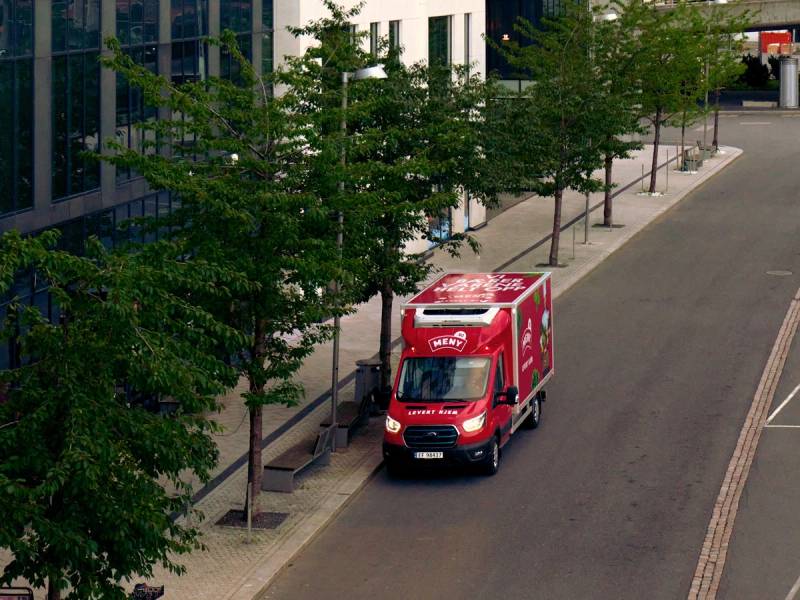 En rød matleveringsbil kjører langs en gate med trær og moderne bygninger.