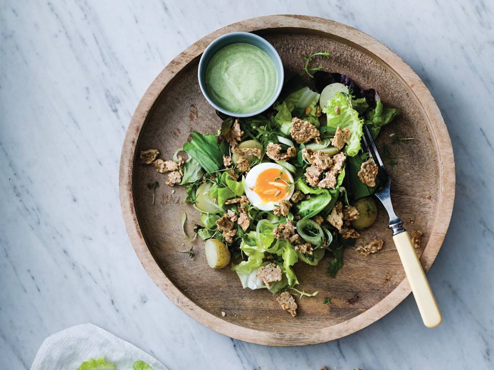 En salat med grønne blader, kokt egg, nøtter og en liten skål med dressing på en rund trefat.