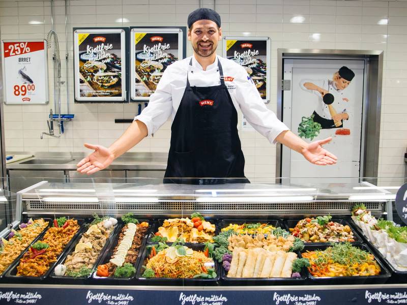 En person i hvit kokkeuniform og svart forkle står bak en salgsdisk med ulike vegetarretter på en matbutikk eller restaurant, med plakater i bakgrunnen.