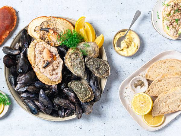 Skål med skalldyr som blåskjell, østers og krabbeklør, sammen med sitronbåter og en liten bolle med smør eller saus, på et lyst bord.