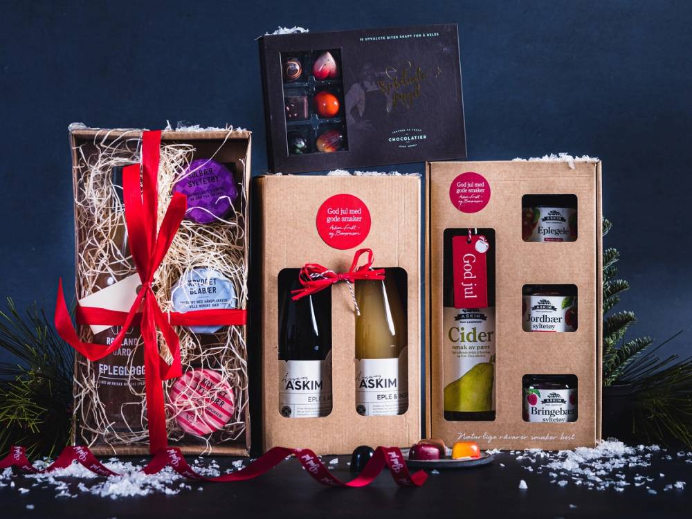 Julegaveesker med matgaver og små flasker, pyntet med rødt bånd og juledekorasjoner, på en mørk bakgrunn.