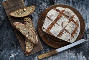 Et brød med grovt innhold og skiver av brød med frø ligger på en treplate, med en kniv ved siden av, på et mørkt trebord.