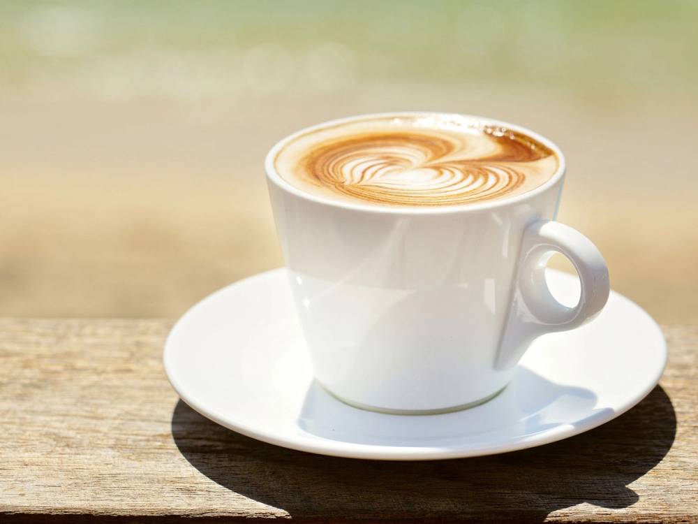 En kopp cappuccino med lattekunst på toppen, plassert på en hvit tallerken på et bord utendørs.