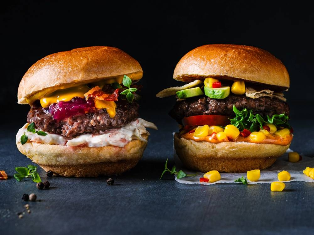 To burgere med ulike fyll, en med ost, salat og kjøtt, og den andre med mais, salat og kjøtt, plassert på en svart bakgrunn.