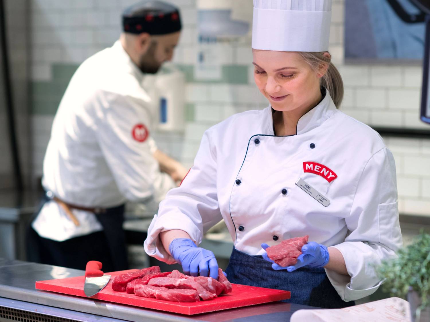 En person i kokkeforkle med blå hansker skjærer kjøtt på en skjærefjøl i en butikk eller restaurant, med en annen person i bakgrunnen.