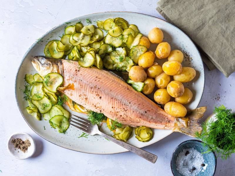 En tallerken med en stekt røye, kokte poteter og agurksalat, med en liten skål med salt og en glass med vann ved siden av.