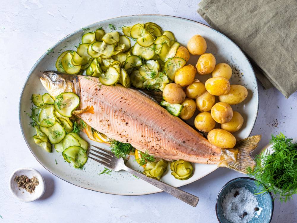 En tallerken med en stekt røye, kokte poteter og agurksalat, med en liten skål med salt og en glass med vann ved siden av.