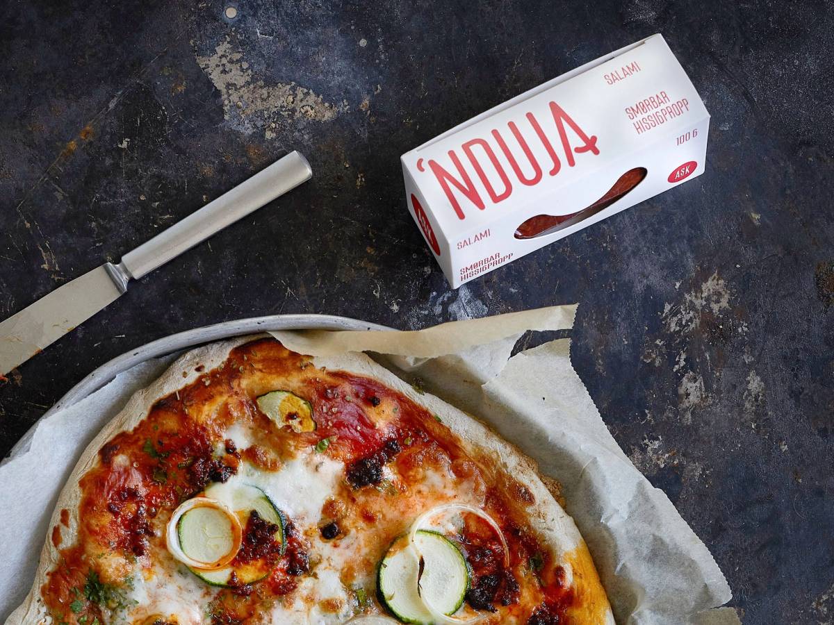 En pizza med skinke, ost og grønnsaker, og en eske nduja ask ved siden av på et mørkt bord.