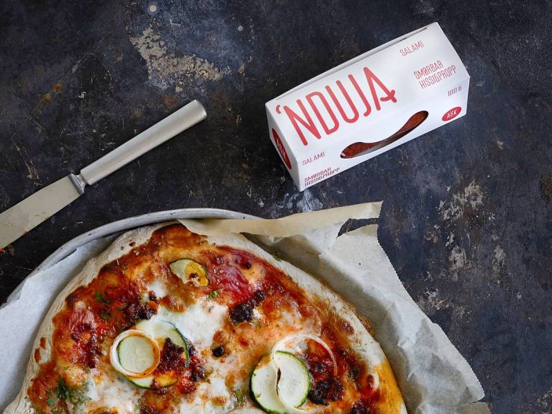 En pizza med skinke, ost og grønnsaker, og en eske nduja ask ved siden av på et mørkt bord.
