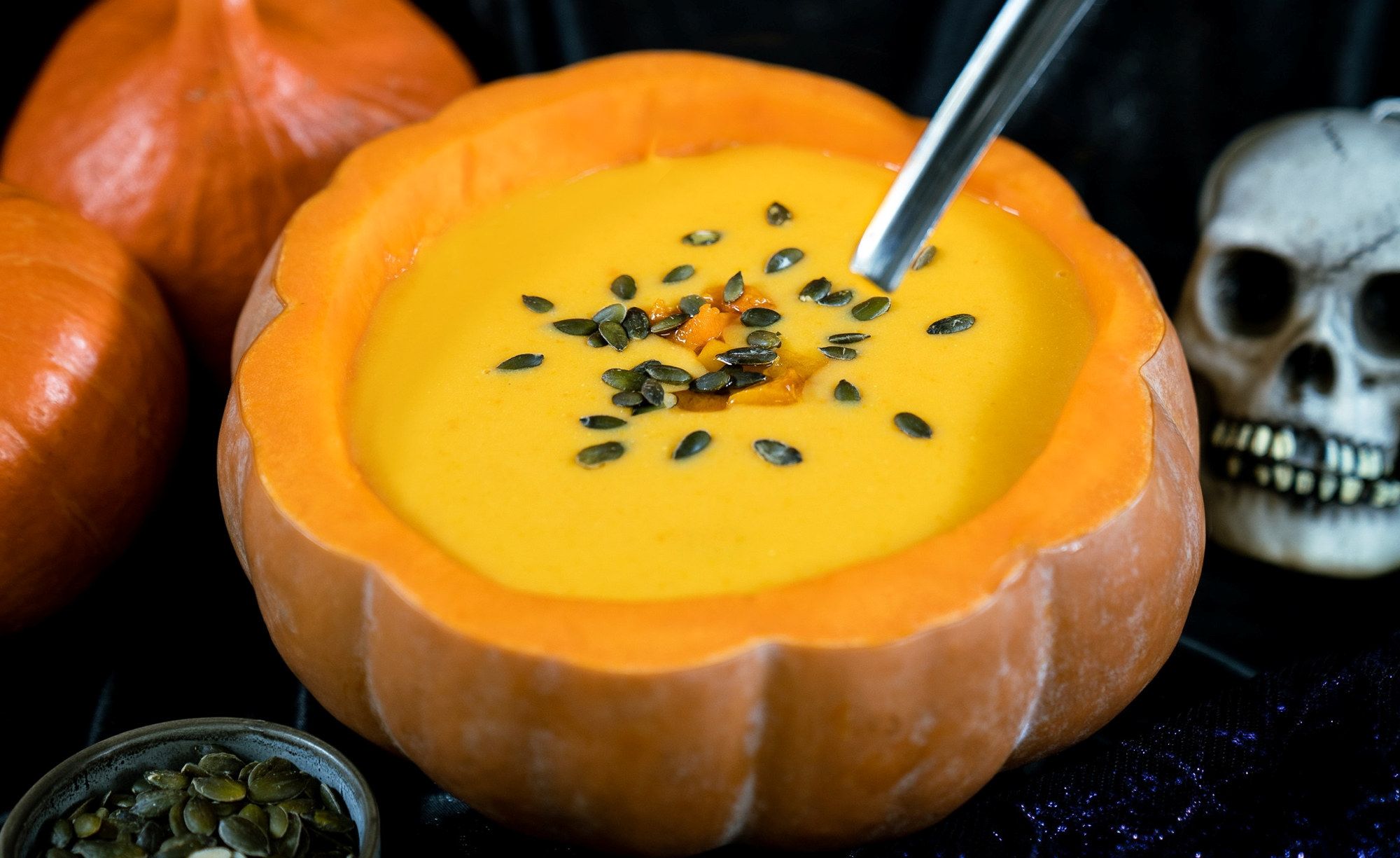 Kraftig gul suppe eller puré i en utstøpt gresskar, pyntet med frø og krydder, omgitt av små gresskar og en skje, med en skjelettskalle i bakgrunnen.