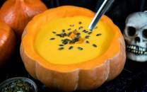 Kraftig gul suppe eller puré i en utstøpt gresskar, pyntet med frø og krydder, omgitt av små gresskar og en skje, med en skjelettskalle i bakgrunnen.