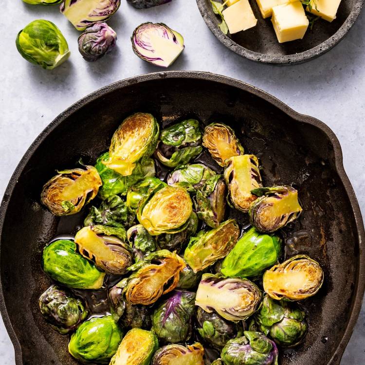 Karamelliserte rosenkål i en svart stekepanne, med noen rå rosenkål og små terninger av ost i bakgrunnen