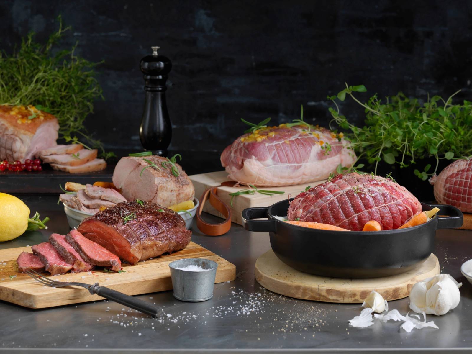 Ulike typer kjøtt, inkludert stek og skiver av rått kjøtt, er presentert på skjærebrett og i en panne på et kjøkkenbord med urter og krydder rundt.