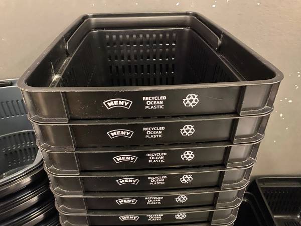 Fem svarte handlekurver merket med 'Recycled Ocean Plastic' og 'Meny' står stablet oppå hverandre mot en vegg.