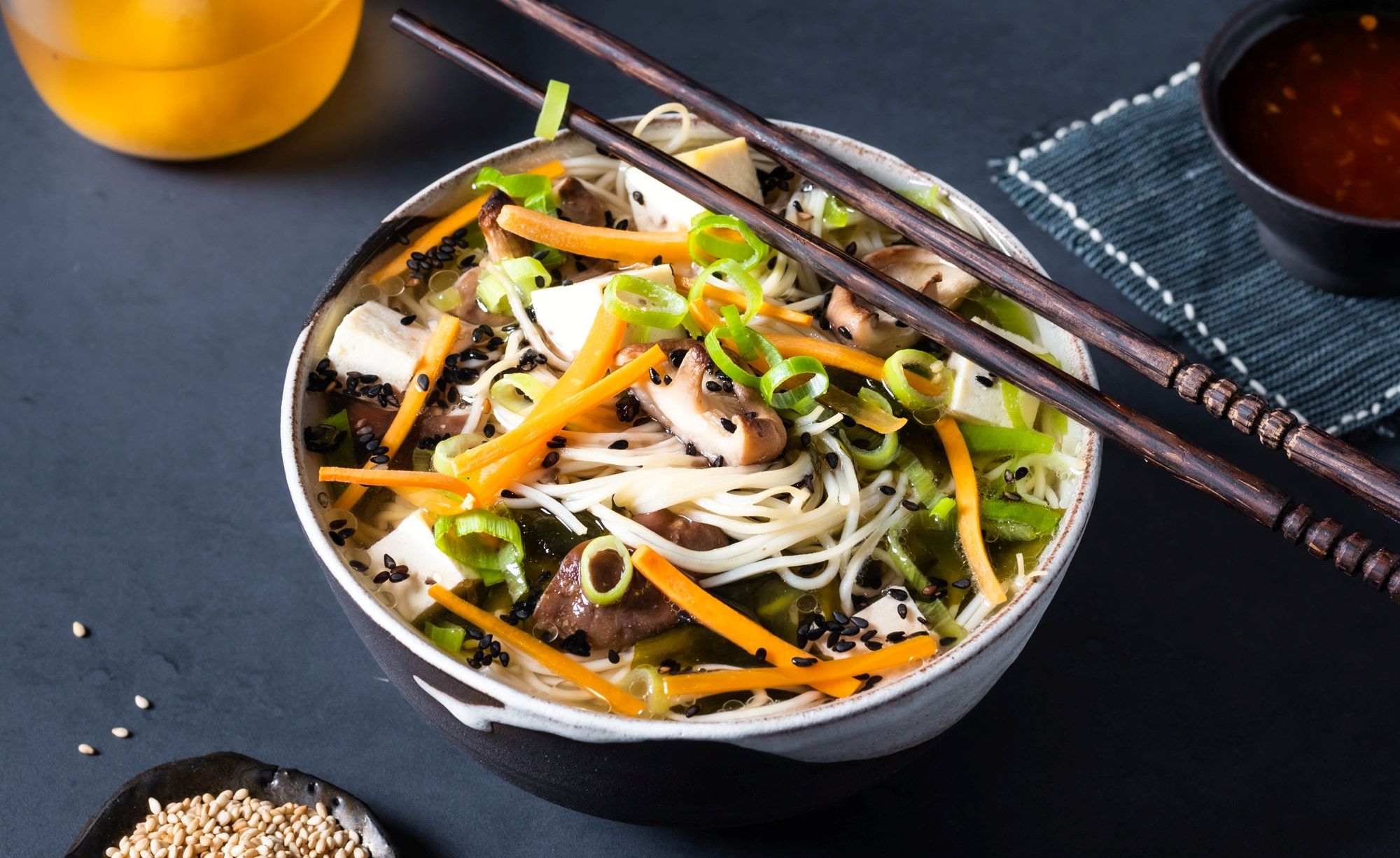 En bolle med nudelsalat med grønnsaker som gulrot, agurk og vårløk, omgitt av spisepinner og en liten skål med saus.