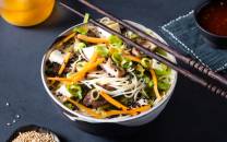 En bolle med nudelsalat med grønnsaker som gulrot, agurk og vårløk, omgitt av spisepinner og en liten skål med saus.