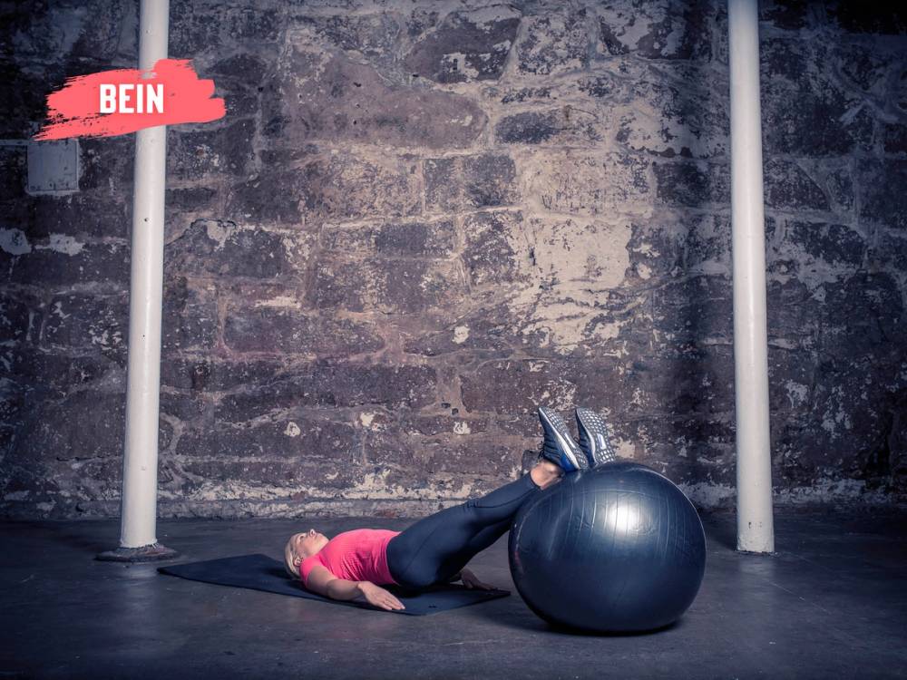 En person ligger på gulvet med beina oppå en stor treningsball i et treningsstudio med murvegg i bakgrunnen.