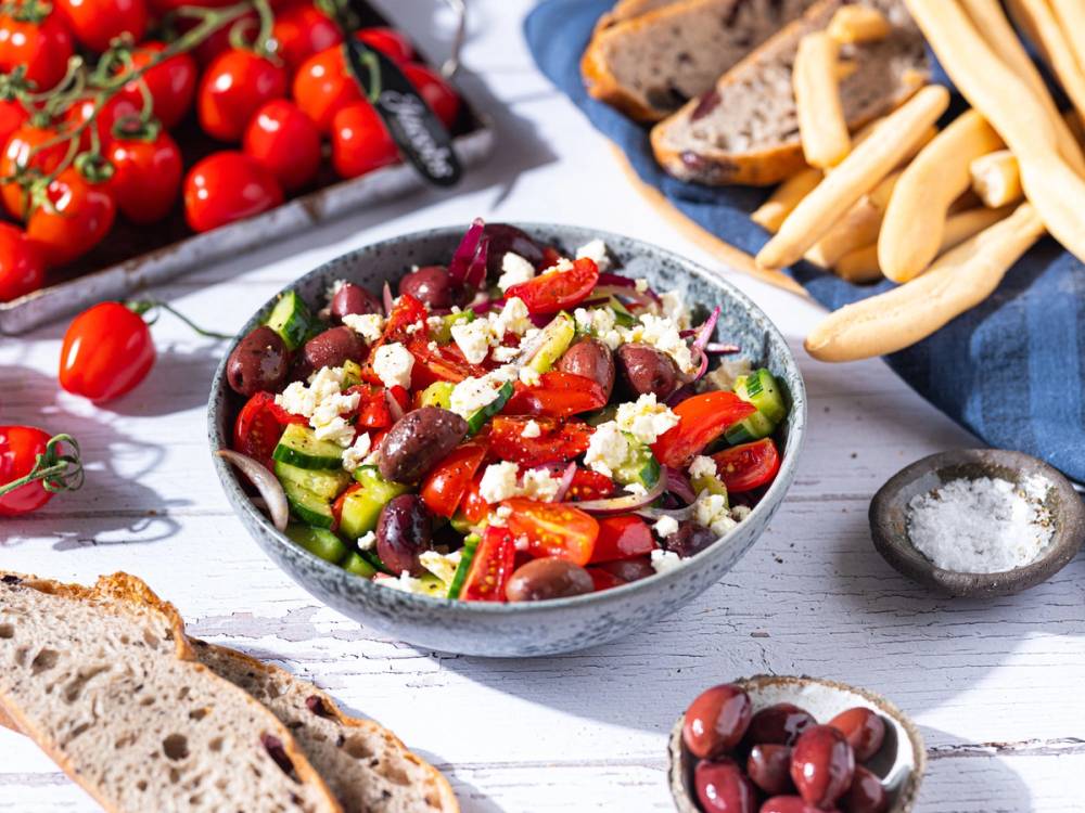 En frisk middelhavs-salat med cherrytomater, oliven, fetaost og grønnsaker i en bolle, omgitt av brød og tomater på et hvitt bord.