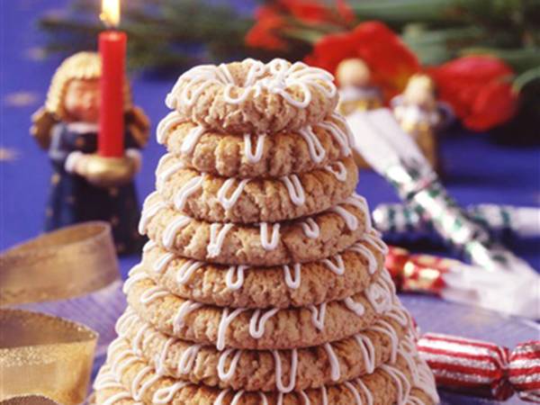 En kransekake dekorert med hvit sjokolade og pyntet med små hvite sjokoladekuler, omgitt av julepynt og lys.