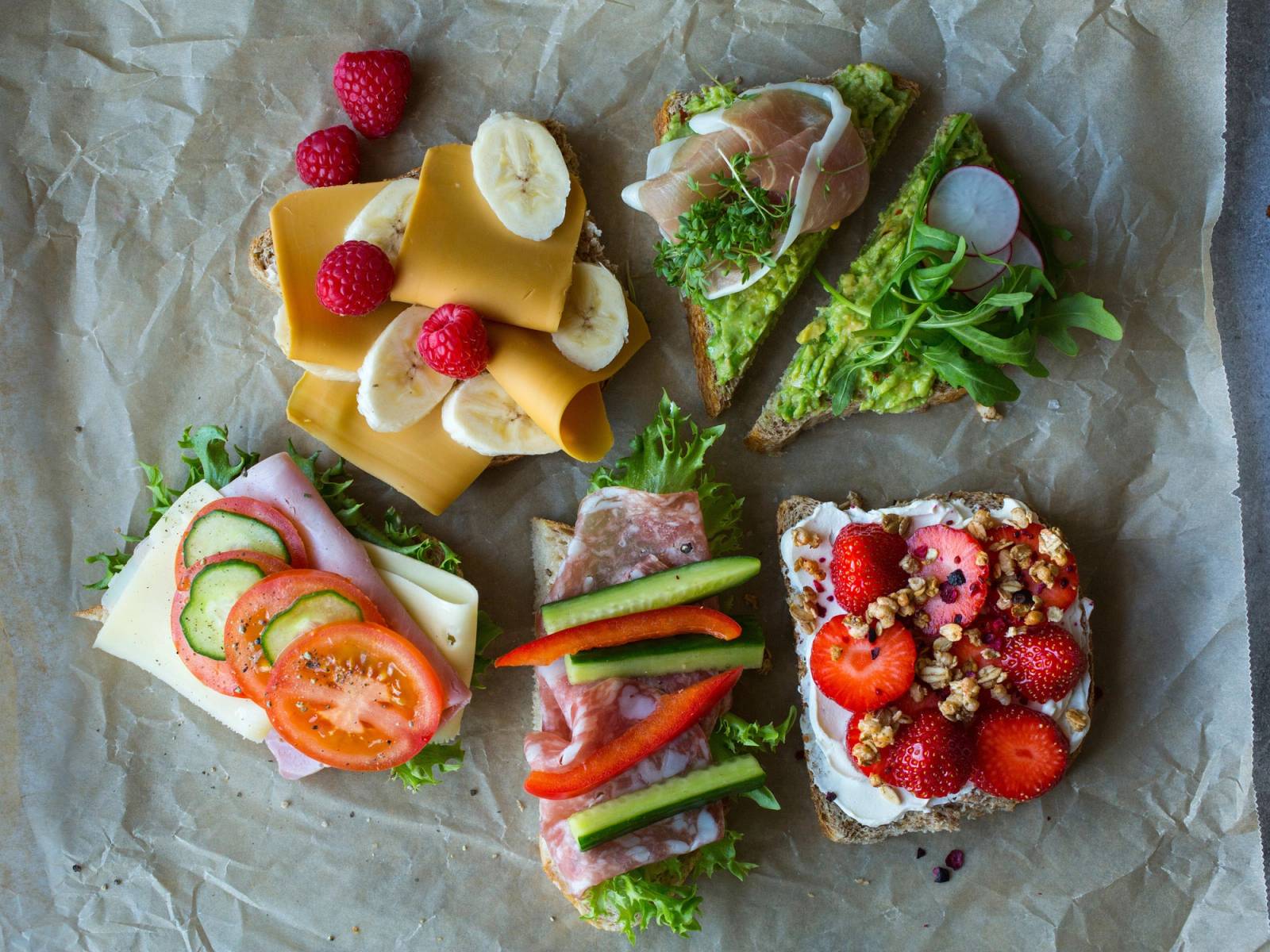 Ulike små matpakker med frukt, grønnsaker og pålegg på papir, klar til skolemat.