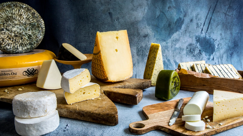 Ulike typer ost og ostekniver på et bord med en blå bakgrunn.