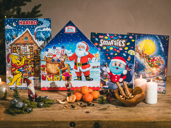 Fem ulike julekalendere står på et bord med julepynt og lys rundt, inkludert en med Haribo-logo og en med Smartees, omgitt av juledekorasjoner og lys.