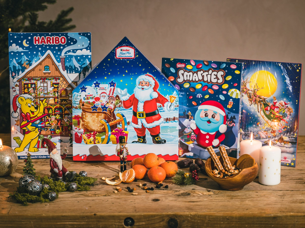Fem ulike julekalendere står på et bord med julepynt og lys rundt, inkludert en med Haribo-logo og en med Smartees, omgitt av juledekorasjoner og lys.