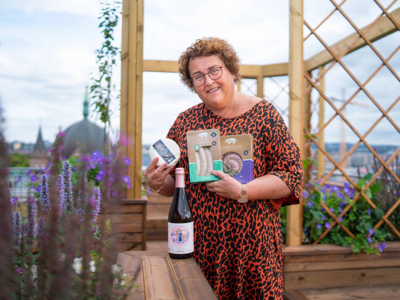 En person holder opp vinnerpremier og en flaske vin på en utendørs terrasse med blomster og treverk i bakgrunnen.