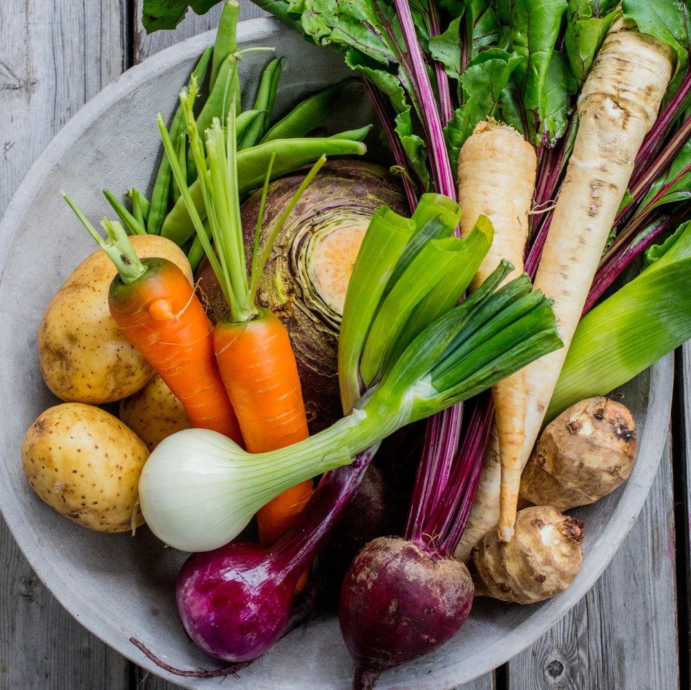 Et utvalg av ferske grønnsaker i en stor bolle, inkludert gulrøtter, purre, poteter, rødbeter, og selleri, på et treverk.