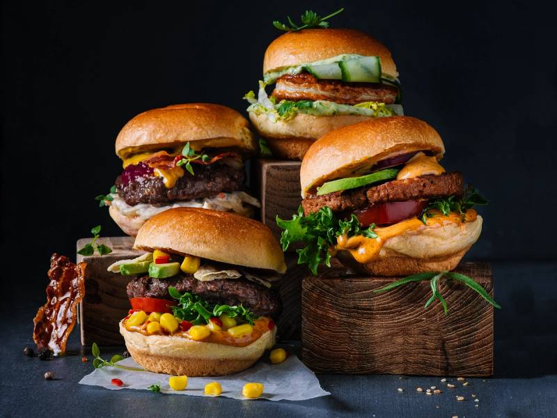 Fire burgere med ulike pålegg og grønnsaker, plassert på og rundt tre trestammer mot en mørk bakgrunn.