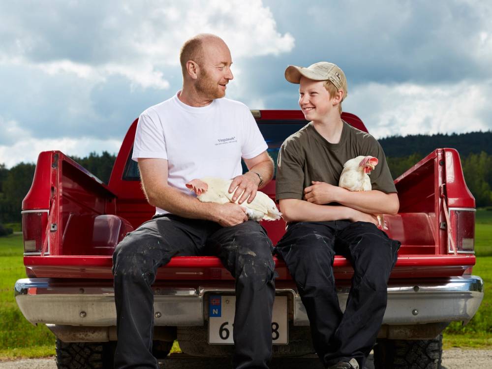 To personer som sitter på en rød pickup, med en åker og skyer i bakgrunnen, og holder på kyllinger.