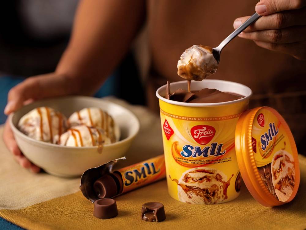 En person spiser iskrem fra en kopp med sjokoladesaus, mens det ligger flere sjokoladebiter og en pakke med Smil-sjokolade på bordet.