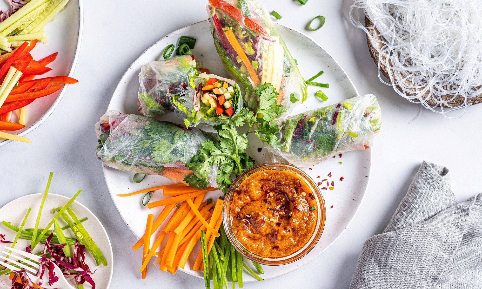 Vegetariske sommerruller med grønnsaker og en liten skål med saus på en hvit tallerken, omgitt av grønnsaker og garnityr.