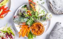 Vegetariske sommerruller med grønnsaker og en liten skål med saus på en hvit tallerken, omgitt av grønnsaker og garnityr.