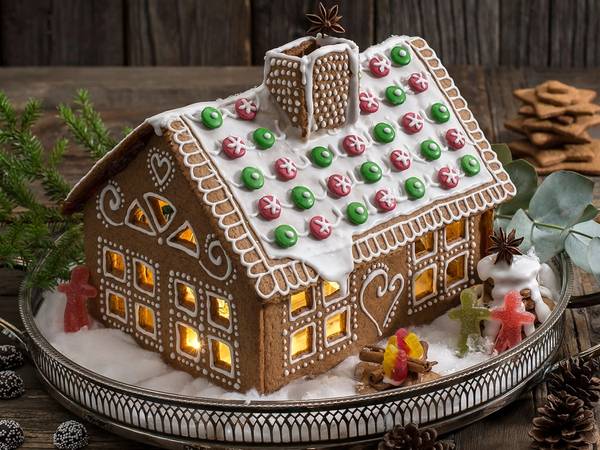 Et pepperkakehus dekorert med fargerike sukkerkuler og lysende vinduer, plassert på et fat med granbar og julekaker rundt.