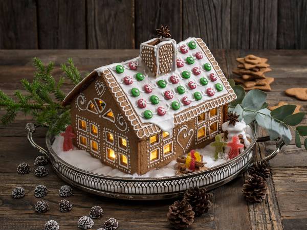 Et pepperkakehus dekorert med fargerike sukkerkuler og lysende vinduer, plassert på et fat med granbar og julekaker rundt.