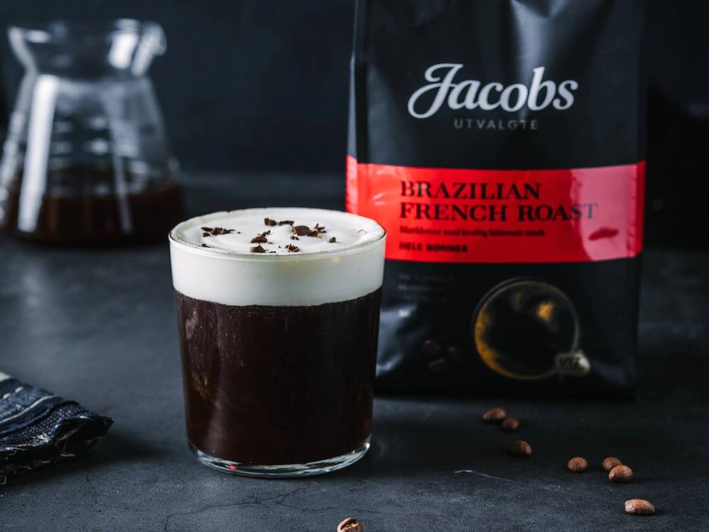 En kopp med julekaffe med krem og sjokoladestrø på toppen, med kaffebønner og kaffepose i bakgrunnen.