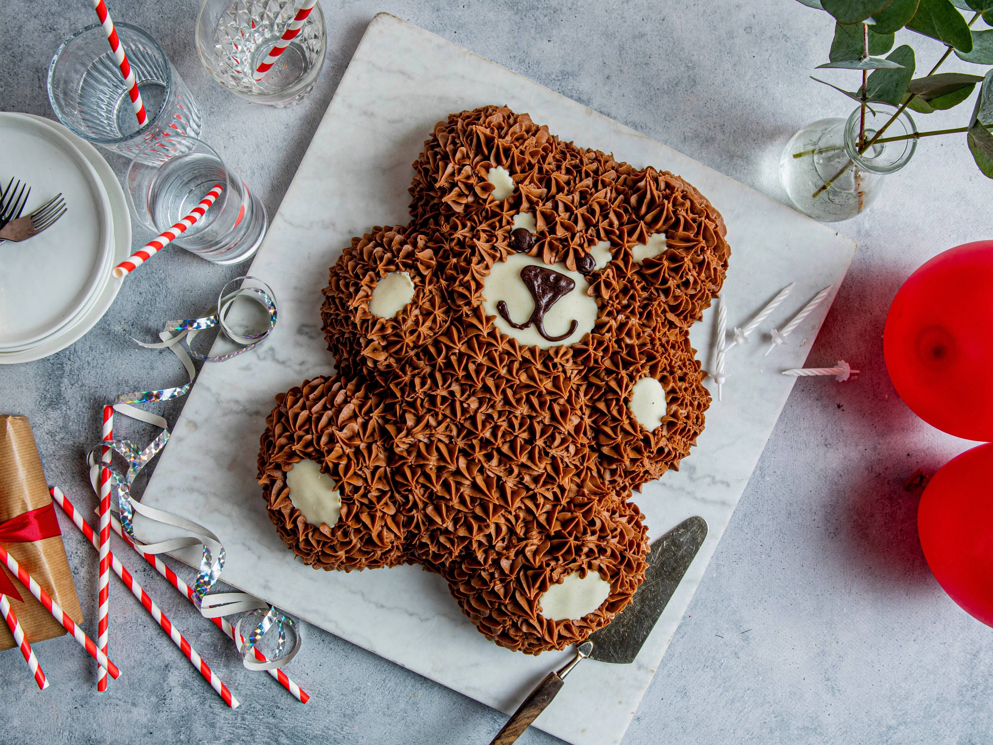 En kake formet som en bamse med sjokoladekrem og hvite detaljer, plassert på et hvitt bord med julepynt rundt.