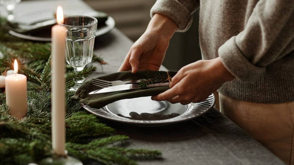 Person som legger på tallerkener på et bord dekket med grønne greiner og lys, sannsynligvis for en julefeiring.