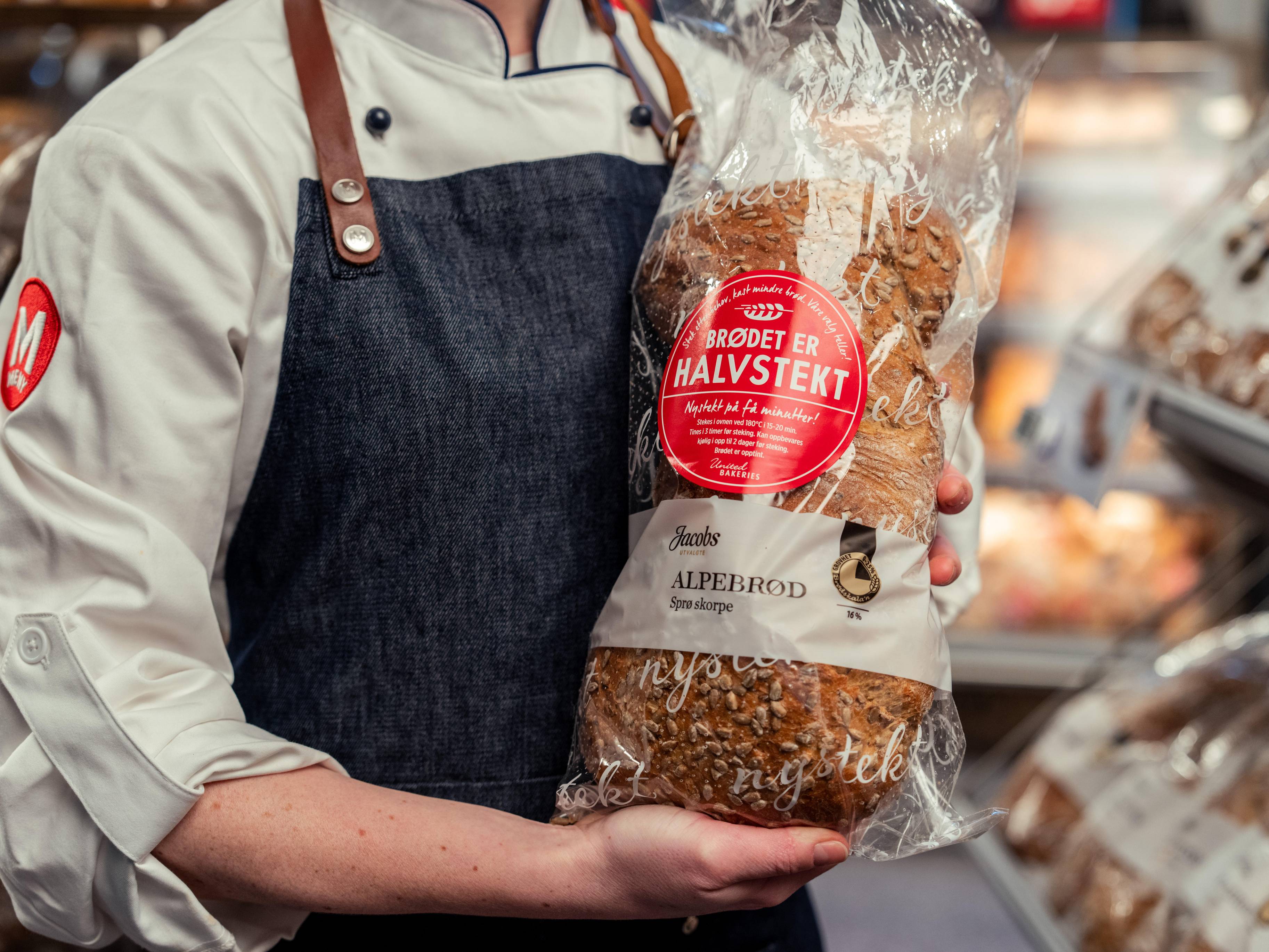 Person som holder en pose med halvstekte brød i en butikk, iført hvit skjorte og mørk apron.