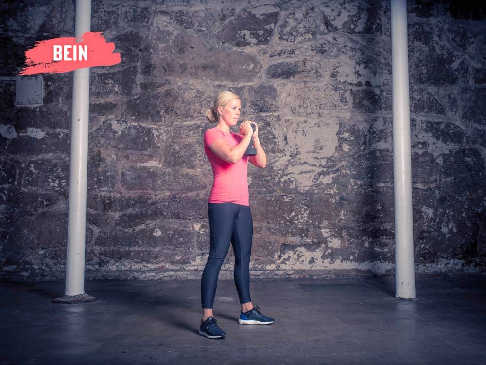 Kvinne utfører en front knebøy med manualer i et treningsstudio med murvegg i bakgrunnen.