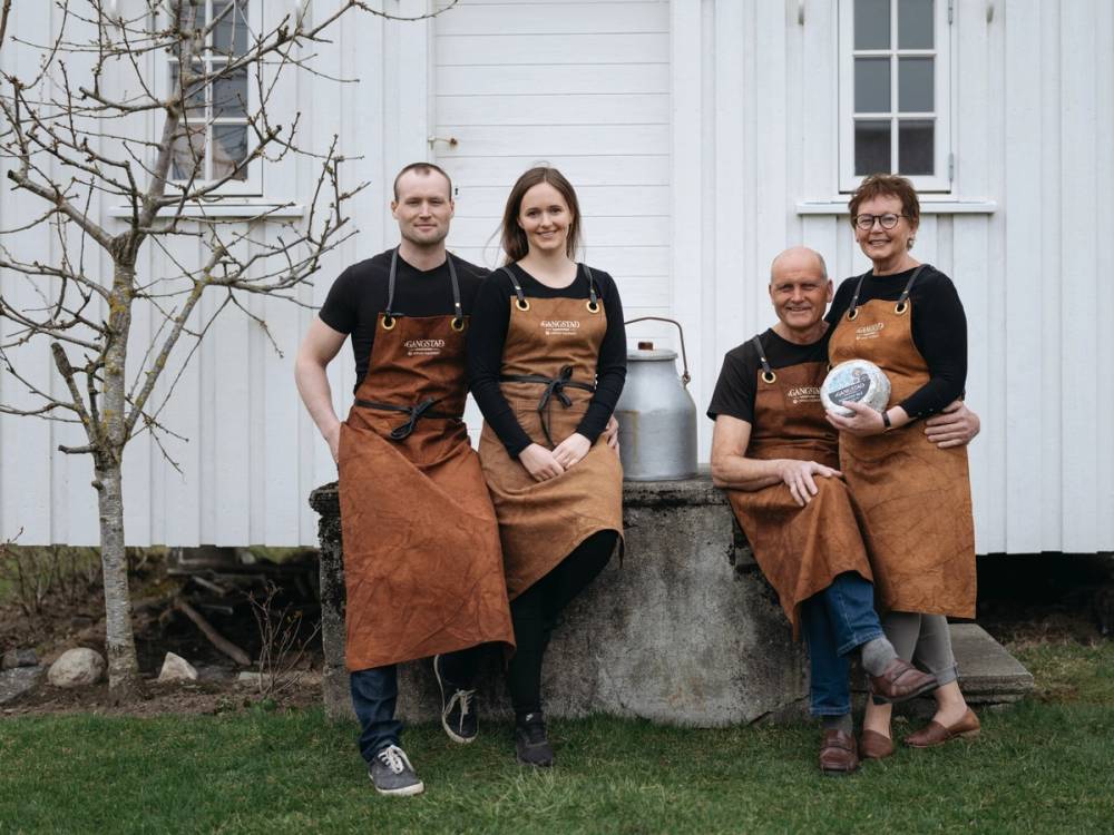Fire personer i brune forkle som sitter og står foran en hvit trebygning, med en melkekanne og en bolle, på en grønn plen.