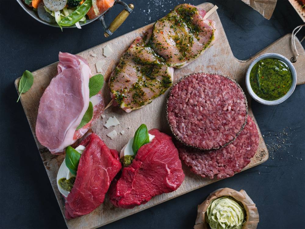 Ulike typer kjøtt, inkludert biff og karbonadedeig, på en skjærebrett med urter og tilbehør, klar for grilling.