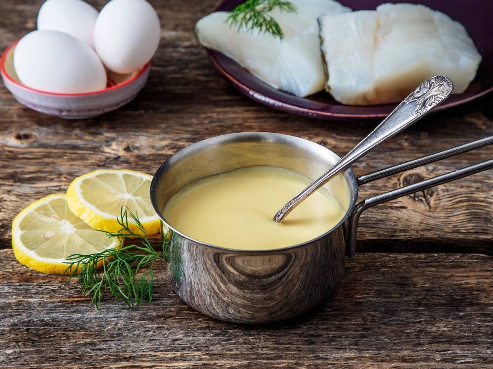 Hollandaise-saus i en liten kjele med sitronbåter og dill ved siden av, samt egg og brød i bakgrunnen.