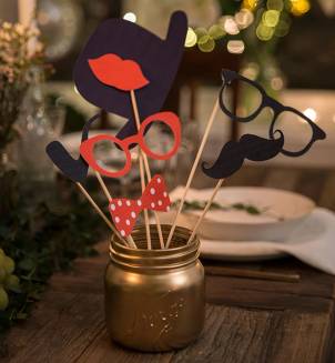 Festlige tilbehør som briller, munn, bart og sløyfe i en gullfarget krukke på et bord, med julelys i bakgrunnen.