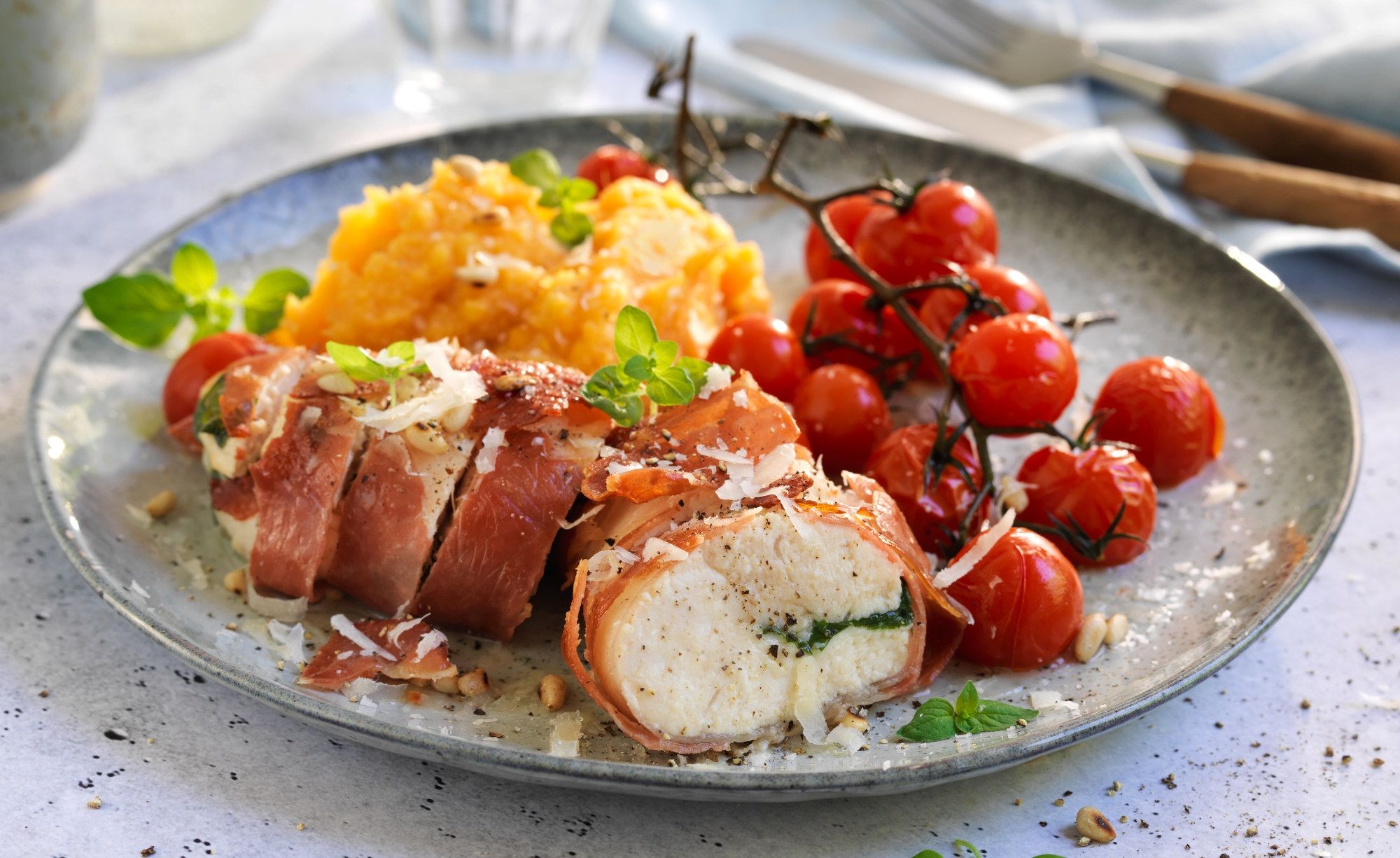 Kyllingbryst fylt med manchago ost og surret med spekeskinke, servert med potetmos og cherrytomater på en tallerken.