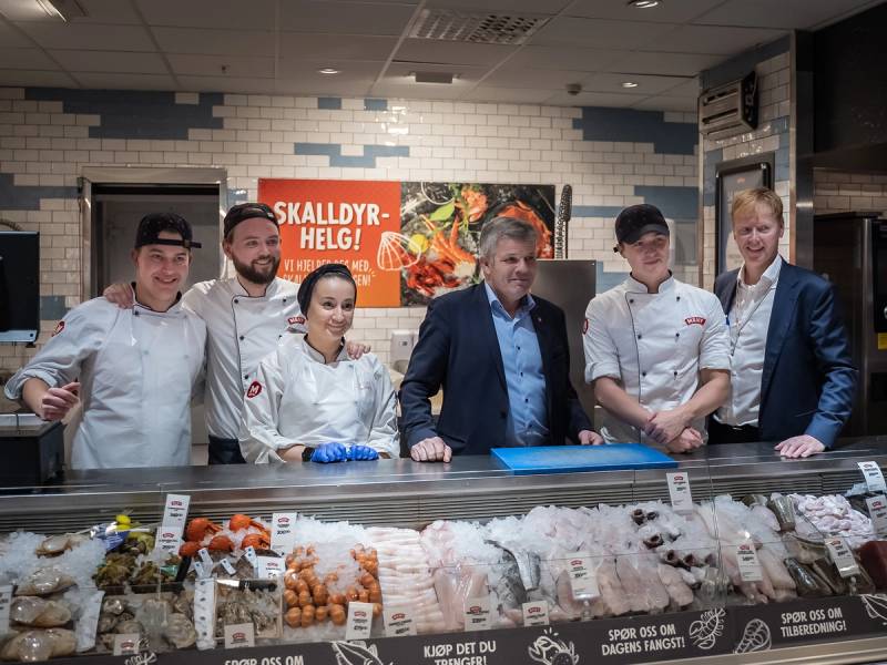 Fiskeriminister står sammen med kokker bak en fiskedisk i en butikk eller marked, med en plakat som sier 'Skalldyrhelg!'.