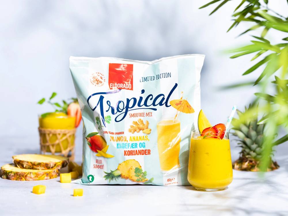 En pose med Eldorado Tropical-snop med smak av mango, ananas, eple og koriander, omgitt av frukt og snacks på et bord.